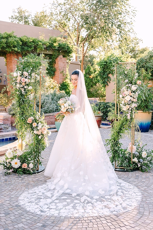 Bridal portrait of a bride in a strapless ball gown with long veil, holding a peach and white bouquet beneath a floral arch in a garden courtyard