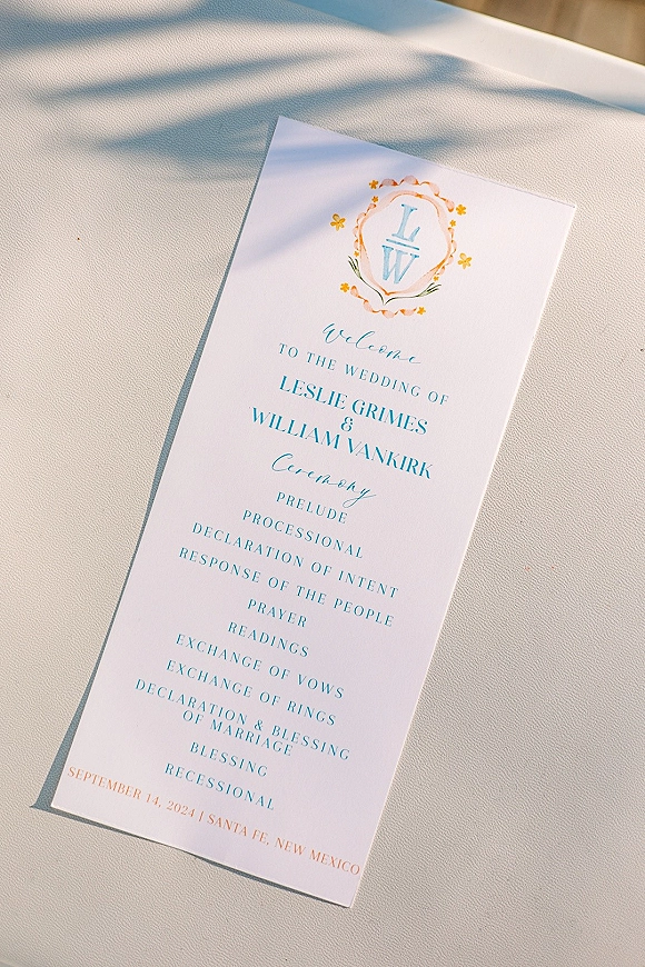 Wedding program ceremony program card with monogram crest and pastel typography on light fabric, dappled by soft sunlight shadows