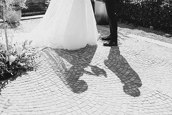 Wedding couple portrait with bride’s ball gown hem and groom’s tuxedo shoes, their shadows cast on a sunlit stone mosaic patio near potted plants