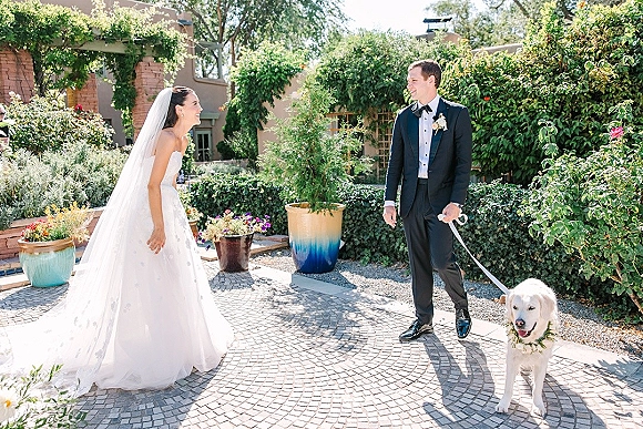 Couple portrait of bride and groom with dog on a leash, laughing in a garden courtyard with brick walls and potted greenery