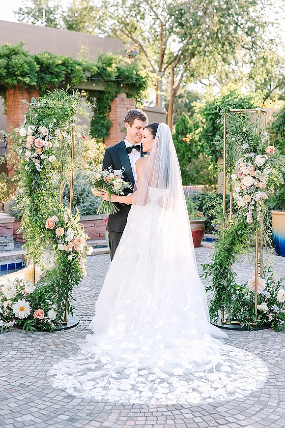 Couple portrait of bride and groom embrace beneath a floral arch, bride holding bouquet with long veil in a garden courtyard