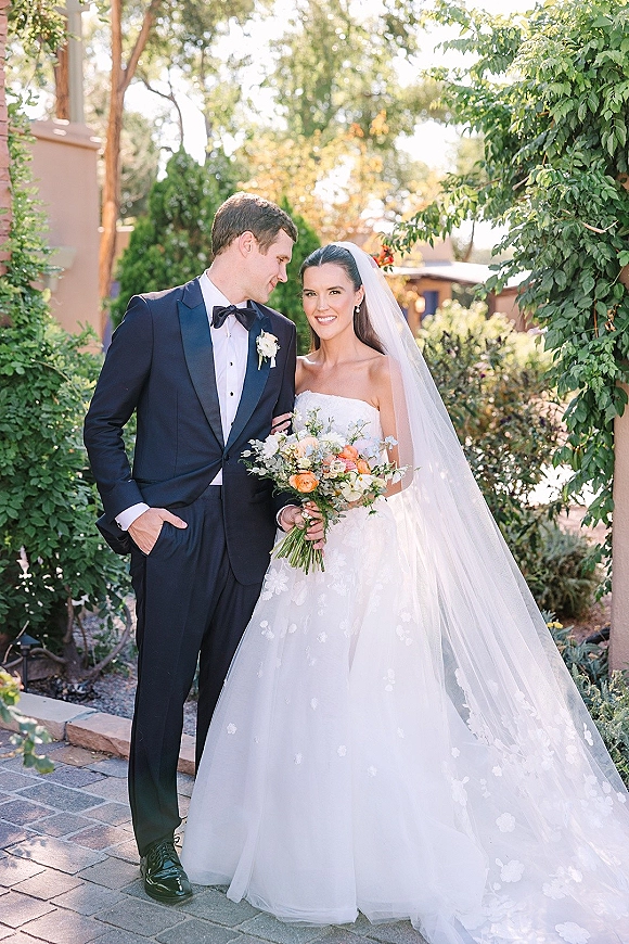 Couple portrait of bride in strapless wedding dress holding a peach and white bouquet beside groom in black tuxedo in a garden courtyard
