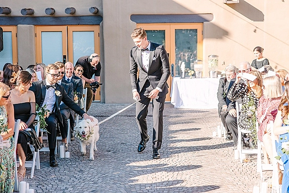 Ceremony processional with groom walking dog down aisle, wearing black tuxedo and boutonniere, past chairs in outdoor courtyard setting.