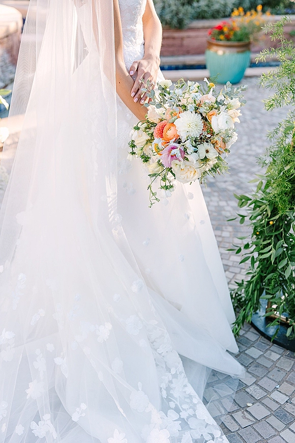 Bridal bouquet of wildflower wedding bouquet blooms in peach, pink, and white with eucalyptus, held over lace dress on a stone patio in a garden setting
