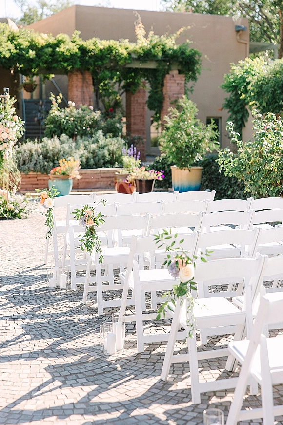 Ceremony seating with white folding chairs lining a floral aisle, greenery garlands and glass cylinder candles in a brick courtyard garden.