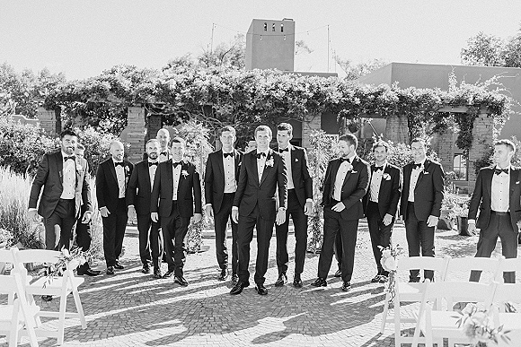 Groomsmen portrait with groom and groomsmen in black tuxedos and bow ties, lined up on brick pavers in a vine-covered courtyard under string lights