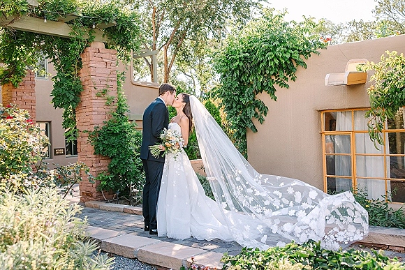 Wedding kiss portrait of bride and groom kissing, her long lace veil trailing and bouquet in hand in a sunlit courtyard garden by stucco wall