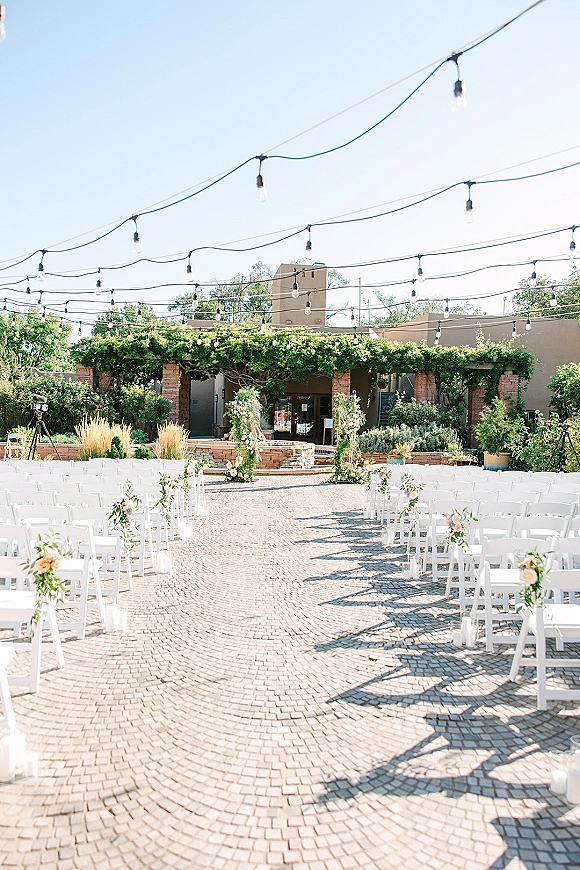 Ceremony setup with outdoor ceremony seating and white folding chairs lining a brick paver aisle to a floral arch under string lights in a courtyard patio