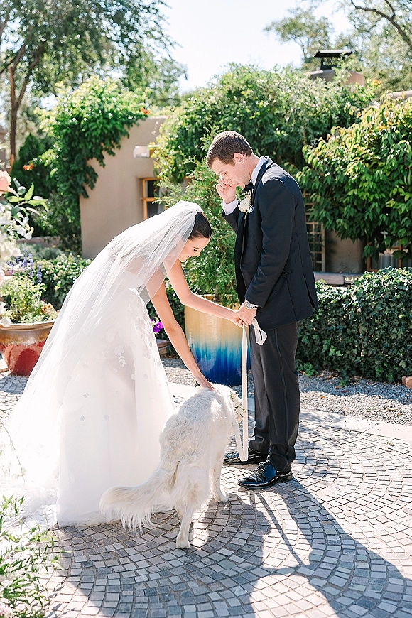 Wedding first look as bride and groom in tuxedo share a tender moment with their dog on leash in a sunlit garden courtyard patio