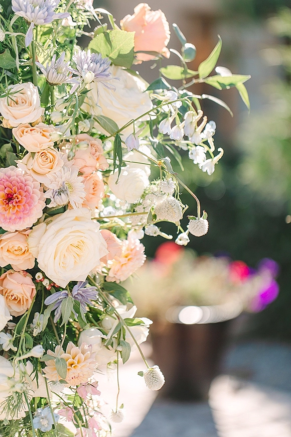 Wedding floral arrangement on a ceremony arch with pastel roses and dahlias, lush greenery, set in an outdoor garden with blurred reception area