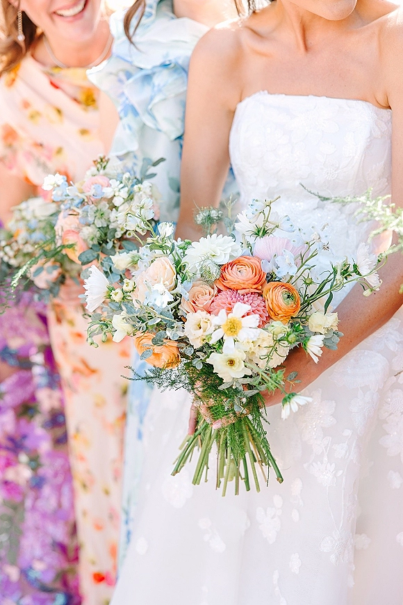 Bridal bouquet, wildflower wedding bouquet in pastel ranunculus, daisies, and blue delphinium held against a strapless lace dress in daylight greenery