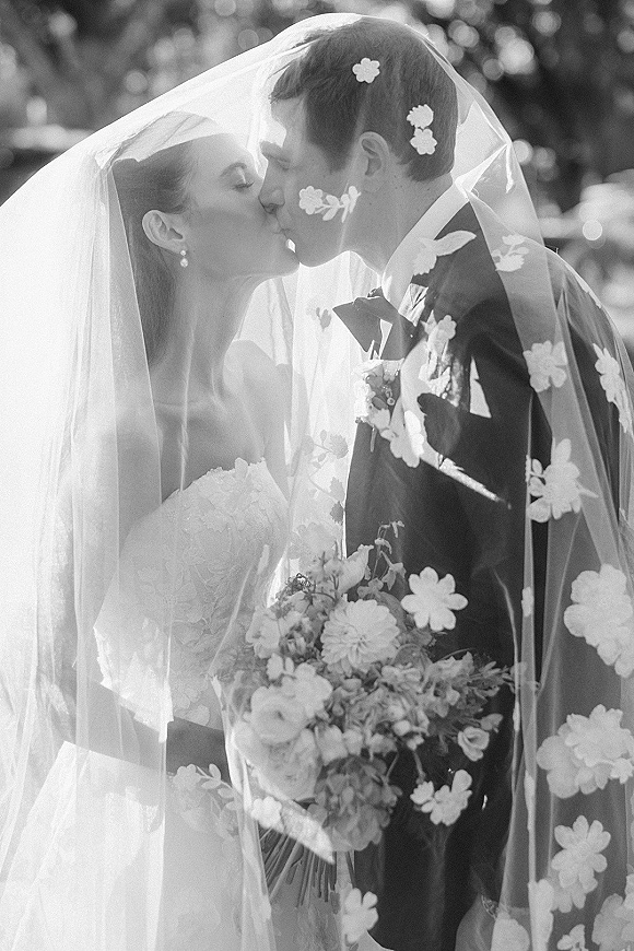 Wedding kiss portrait of bride and groom kissing under a floral appliqué veil, bouquet in hand, with sunlit trees in soft bokeh behind