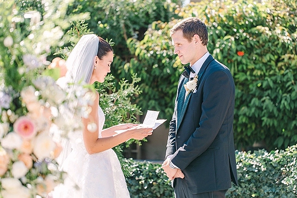 Wedding vows as bride reading vows to groom in a tuxedo, holding vow cards, with a bridal veil in sunlit garden greenery backdrop