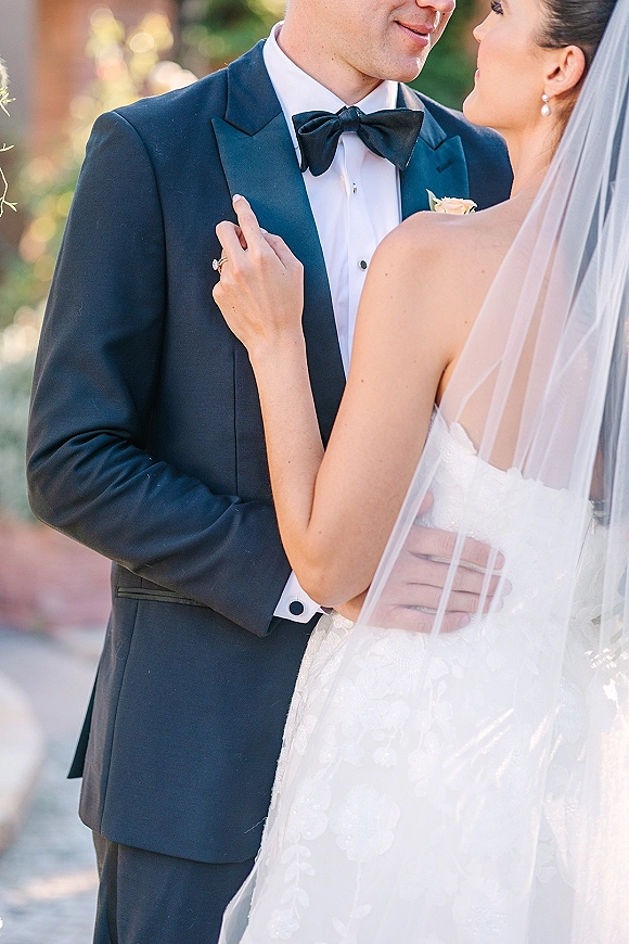Couple portrait of bride and groom embrace, her hand on his chest showing engagement ring, veil and tuxedo against greenery and brick bokeh lights