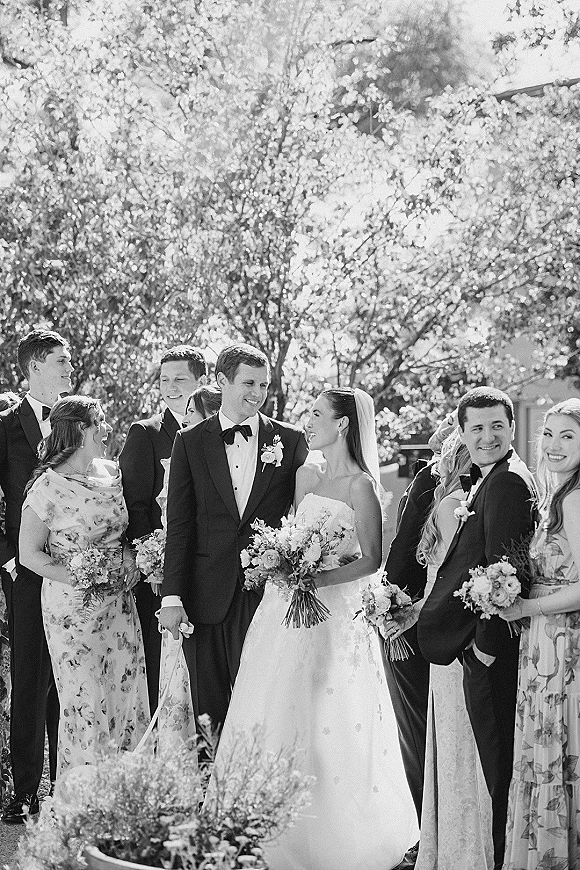 Wedding party portrait with bride and groom with wedding party, bride in veil holding bouquet beside tuxedoed friends in a sunny garden