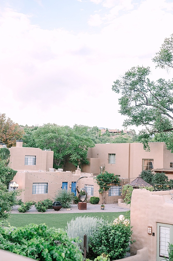 Outdoor venue courtyard with courtyard string lights above a lawn, surrounded by stucco buildings, trees, and a sculpture at dusk