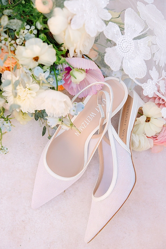 Bridal shoes, white wedding heels in glitter slingback pointed-toe style beside a bouquet and lace veil on light stone surface