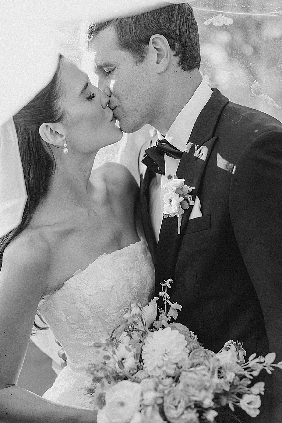 Wedding kiss in a black and white wedding photo, bride in strapless lace gown and veil kissing groom in tux amid sunlit greenery bokeh