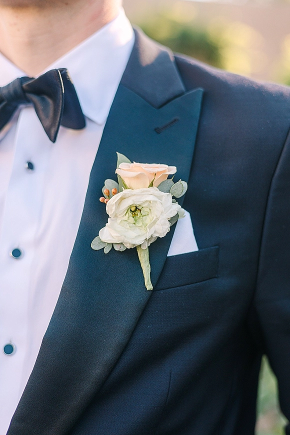 Groom boutonniere with a white and peach rose wedding boutonniere pinned to a navy tuxedo lapel, set against blurred outdoor greenery