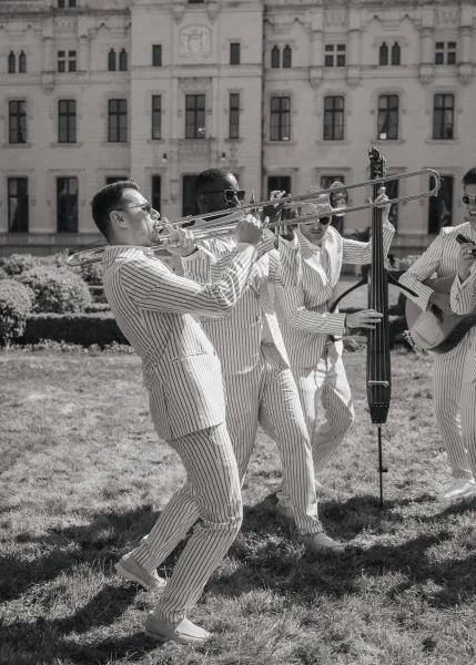 wedding musicians performing as a live wedding band on a lawn, featuring a trombone, with hedges and a stone building facade behind them