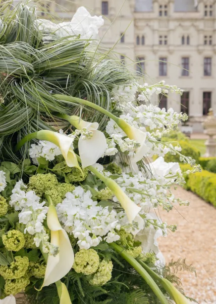 Ceremony flowers in a calla lily wedding arrangement, framed by garden hedges along a gravel path before a historic building facade.