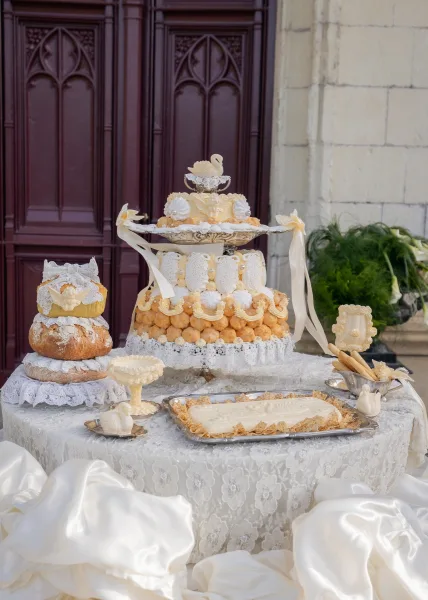 Wedding dessert table featuring a croquembouche wedding cake, set before carved wooden doors and a stone wall with greenery.