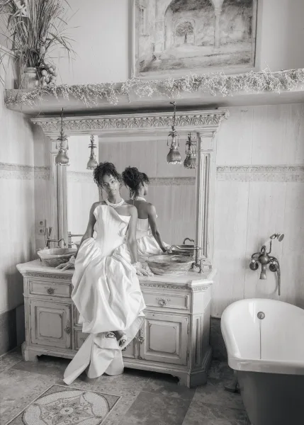 Bridal portrait of a bride in a strapless gown, wearing a pearl necklace, seated in a tiled bathroom with stone floor and wall art.