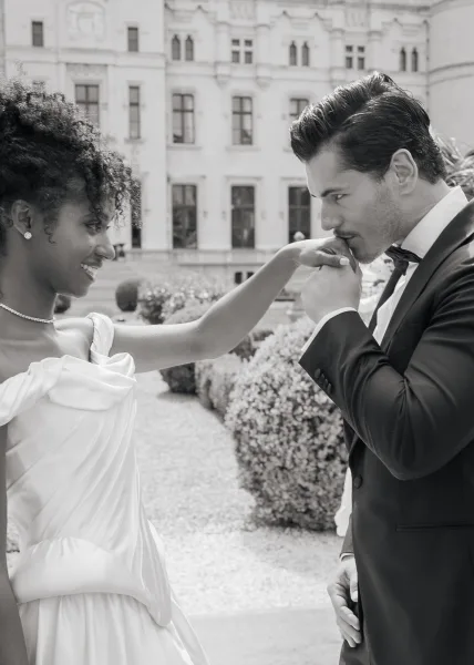 Couple portrait with the groom kissing the bride's hand, the bride wearing a pearl necklace, in a formal garden amid topiaries by a historic facade.