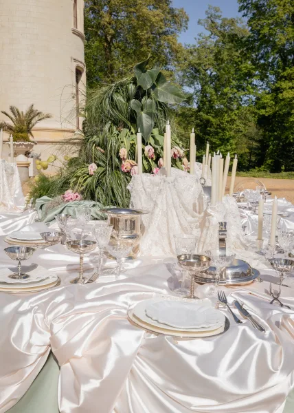 Reception tablescape atop an outdoor reception table with glowing taper candles, framed by garden trees and a stone building beneath blue sky.