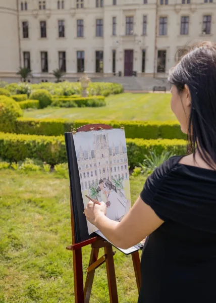 Live wedding painting by a wedding painter at an easel, paintbrush poised, capturing the bride and groom in a formal garden before a stone facade