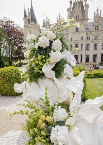 Wedding floral installation of white and green wedding flowers with white hydrangeas frames a castle facade along a gravel path.