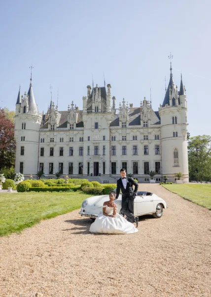 Couple portrait captured as an outdoor wedding portrait beside a vintage convertible, framed by a castle facade and formal gardens.