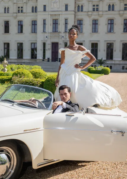 Couple portrait of a bride and groom with a vintage convertible on a gravel driveway before a grand mansion facade and formal garden.