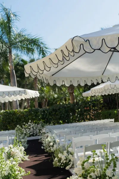 Outdoor ceremony setup with wedding ceremony aisle lined by white folding chairs and low white florals, palm trees and umbrellas under blue sky