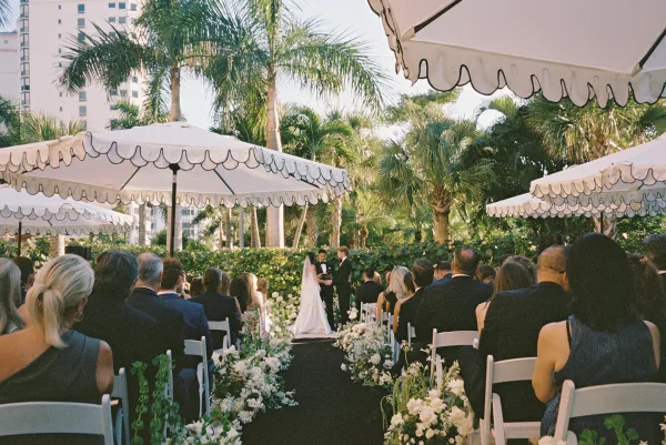 Wedding ceremony with bride and groom at altar, guests under scalloped white umbrellas beside floral-lined aisle and palm trees