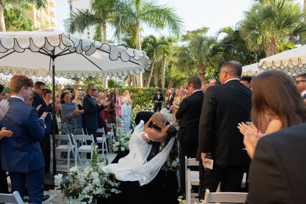 Ceremony kiss as bride dips in veil and strapless dress, groom in tuxedo, guests cheering along a flower-lined aisle with palms behind
