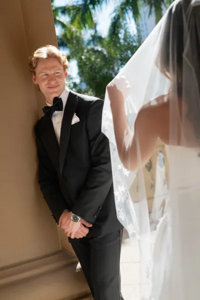 First look moment as bride in a strapless dress and veil taps groom in tuxedo on a sunlit palm-lined walkway by columns