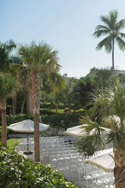Outdoor ceremony setup with beach wedding ceremony chairs in neat rows, white umbrellas for shade, and floral arrangements amid palms under blue sky