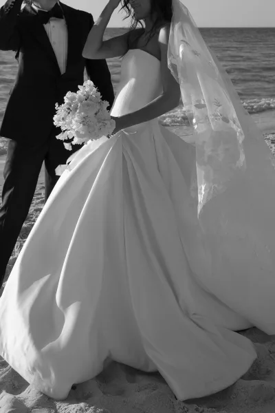 Couple portrait on a beach wedding portrait, bride in strapless gown holding bouquet beside groom in tuxedo with ocean waves behind