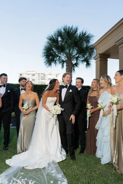 Wedding party portrait of bride and groom with bridesmaids laughing beside groomsmen in tuxedos, with palm tree and columns behind