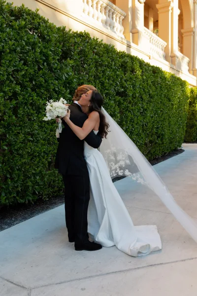 Wedding couple portrait of bride and groom hug, her long veil trailing as she holds a white bouquet by green hedge and columns outdoors