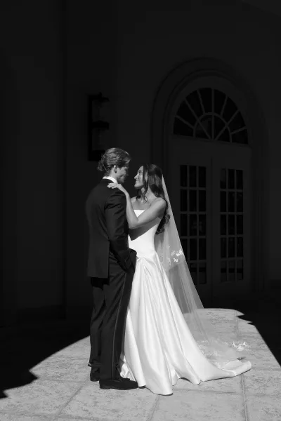 Couple portrait in a black and white wedding portrait style, bride in strapless dress and veil embracing groom by a sunlit arched doorway