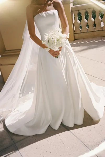 Bridal portrait of a bride in a strapless wedding dress holding a white bouquet, cathedral veil trailing on a sunlit stone terrace