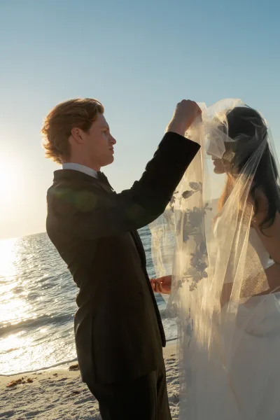First look moment as groom lifting veil from bride in strapless gown, tuxedo and bow tie on a beach with ocean behind