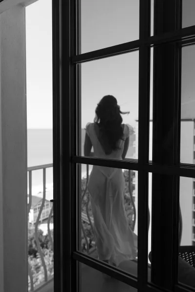 Bridal portrait of a bride at window in an open-back gown with flutter sleeves, long hair, on a balcony overlooking the ocean and sky