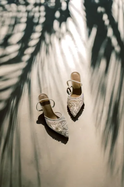 Wedding shoes with pearl beading, white pointed toes and ankle straps in a sunlit flatlay with palm leaf shadows on the floor