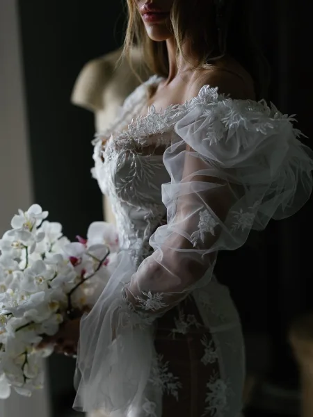 Wedding dress with off the shoulder wedding dress lace appliqué and sheer tulle sleeves, held with white orchid bouquet in dark window light