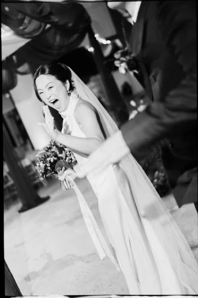 Bride portrait of a laughing bride holding bouquet, waving in an indoor reception with guests and soft bokeh lights behind her