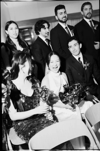 Wedding party portrait of a bride laughing with bridesmaids and groomsmen in suits, seated on chairs in a wood-paneled indoor venue