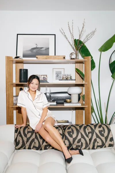 Bridal getting ready bride in pajamas sits on a sofa in a white living room, showing earrings and engagement ring in satin set